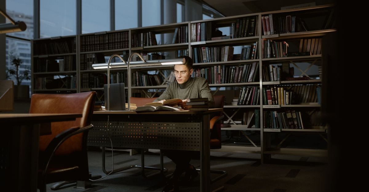 Person studying at a desk in a library between tall bookshelves