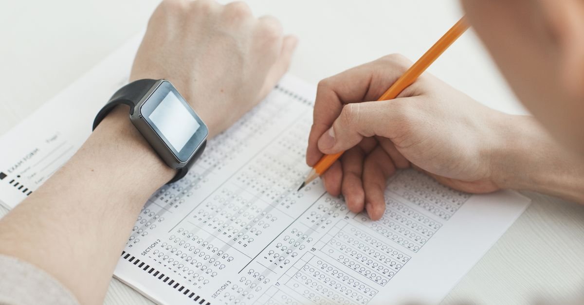 Hand filling in an answer sheet with a pencil beside a watch