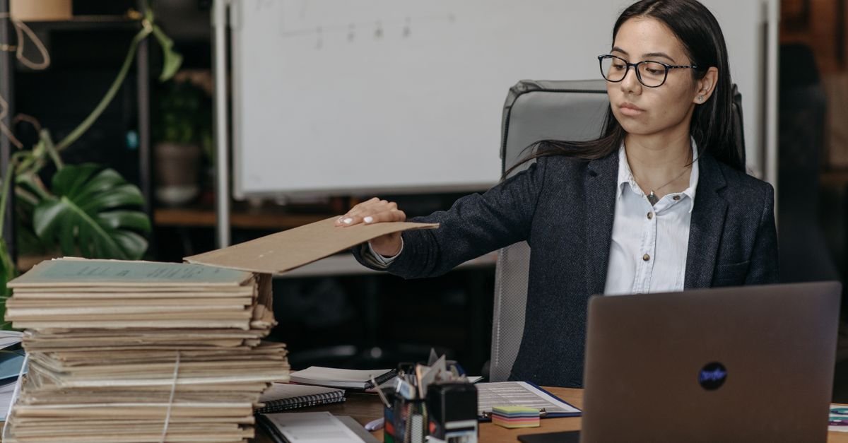 Office worker beside tall stacks of paper files, reaching for a folder in a licensing queue